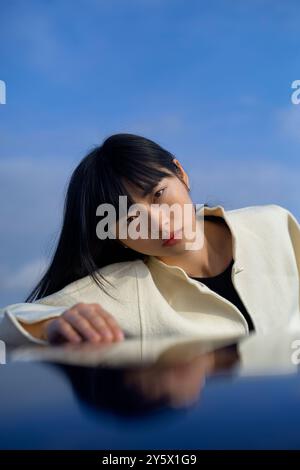 Porträt einer nachdenklichen jungen Frau mit schwarzen Haaren, die ihren Kopf auf dem Arm vor einem blauen Himmel auflegen. Stockfoto