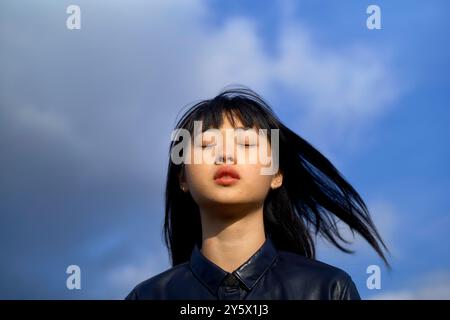 Nahaufnahme einer jungen Frau mit schwarzem Haar und Augen geschlossen vor blauem Himmel mit Wolken. Stockfoto