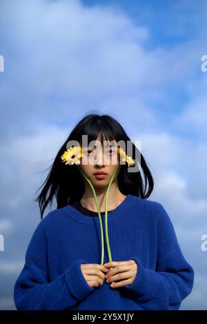 Eine heitere junge Frau, die gelbe Blumen vor einem wolkigen blauen Himmel hält. Stockfoto