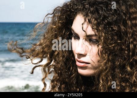 Lockige Frau, die nachdenklich mit dem Meer im Hintergrund blickt. Stockfoto