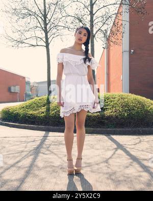 Eine Frau in einem weißen schulterfreien Kleid steht auf einem Bürgersteig mit Bäumen und einem Backsteingebäude im Hintergrund. Stockfoto