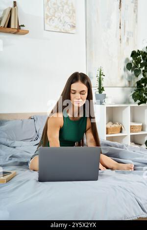 Eine junge, brünette Frau mit kurzer Größe sitzt bequem auf ihrem Bett, beschäftigt sich mit ihrem Laptop, während sie ihre Zeit zu Hause genießt. Stockfoto