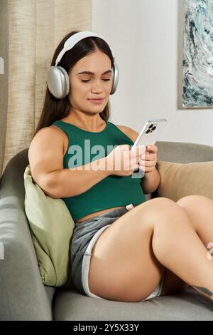 Eine kurzstattige Frau entspannt sich auf einer Couch, die durch ihre Kopfhörer in Musik eingetaucht ist, und genießt ihren gemütlichen Raum. Stockfoto