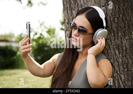 Eine junge, brünette Frau mit kurzer Größe entspannt sich draußen, trägt Kopfhörer und macht ein Selfie, um den Moment zu genießen. Stockfoto