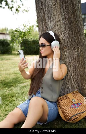 Eine junge, brünette Frau mit kurzer Größe entspannt sich draußen, sitzt an einem Baum, hört Musik und überprüft ihr Telefon. Stockfoto