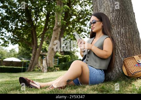 Eine junge Frau mit kurzer Größe genießt einen hellen Tag im Freien, sitzt unter einem Baum und greift sich mit ihrem Smartphone an, während sie sich entspannt. Stockfoto