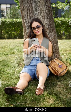 Eine junge Frau mit brünetten Haaren entspannt sich in der Sonne, sitzt unter einem Baum, während sie sich auf ihr Smartphone konzentriert, und nimmt den Moment wahr. Stockfoto