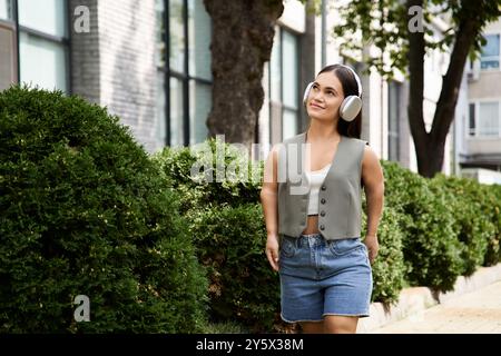 Eine kleine junge Frau mit kurzer Größe schlendert im Freien, trägt Kopfhörer und genießt die Wärme eines sonnigen Tages. Stockfoto