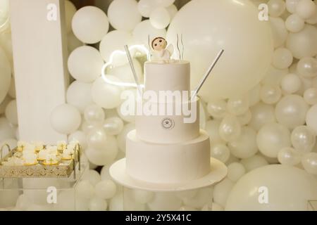 Ein wunderschön dekorierter weißer dreistöckiger Kuchen mit einer Engelsfigur auf der Oberseite, umgeben von weißen Ballons, perfekt für eine Feier. Stockfoto