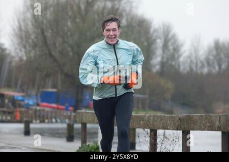 Ein lächelnder Mann, der im Regen joggt, trägt eine hellblaue Jacke, eine schwarze Hose und orangefarbene Handschuhe. Stockfoto