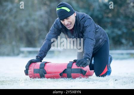 Mann in Wintersportbekleidung grimmend, während er einen schweren roten Sandsack während eines Outdoor-Workouts auf einem frostigen Feld aufnimmt. Stockfoto