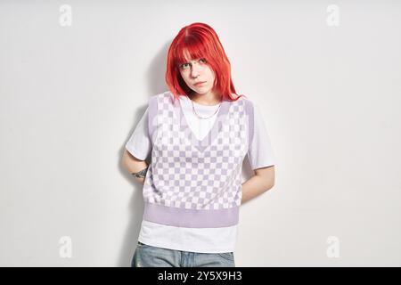 Porträt einer jungen rothaarigen Frau, die vor weißem Hintergrund steht, während sie direkt in die Kamera blickt, in karierter Weste und lässigem Outfit mit Händen auf der Hüfte Stockfoto