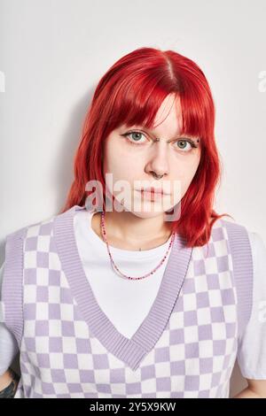 Porträt einer jungen Frau mit leuchtend roten Haaren, die eine karierte lila Weste über weißem Hemd und Perlenkette trägt, direkt vor der Kamera mit neutralem Ausdruck Stockfoto