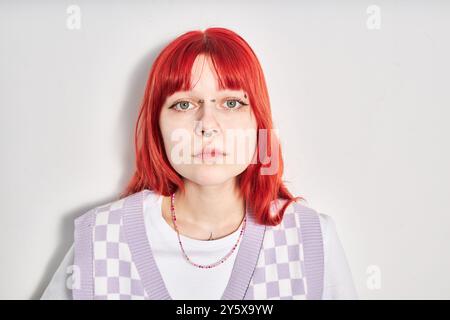 Porträt einer jungen Frau mit hellrotem Haar und Piercing, die lila karierte Weste und weißes Hemd trägt, an der weißen Wand steht und direkt in die Kamera blickt Stockfoto