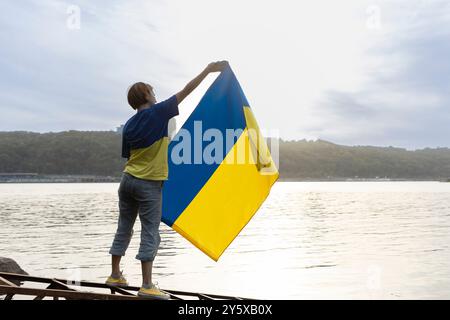 Frau steht vor dem Hintergrund eines Flusses und hält eine ukrainische gelb-blaue Flagge. Stockfoto