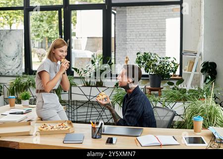 Zwei Kollegen teilen sich einen fröhlichen Moment bei einer Pizza, während sie im Büro von Grün umgeben sind. Stockfoto