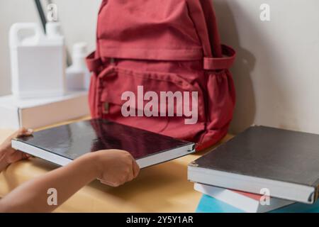 Ein Schüler organisiert einen Schreibtisch, indem er einen roten Rucksack mit Lehrbüchern packt und sich auf die Schule oder das Studium vorbereitet. Stockfoto
