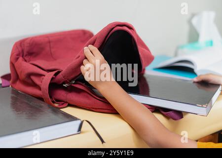 Ein Kind packt Schulbücher in einen roten Rucksack auf einem Schreibtisch und bereitet sich auf die Schule vor. Die Szene vermittelt Organisation und Bereitschaft und betont Bildung und Stockfoto