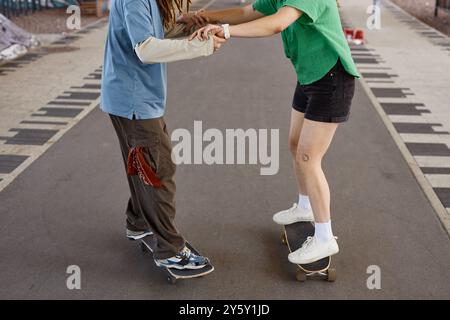 Zwei Personen lernen Skateboarden, während sie Hände halten und auf einem asphaltierten Pfad im Park stehen. Eine Person hat hochgerollte Ärmel und beide tragen legere Kleidung Stockfoto