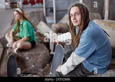 Porträt eines jungen Mannes mit Dreadlocks, der Skateboard hält, während er auf Felsen in der Stadt sitzt. Frau im Hintergrund sitzend mit überkreuzten Beinen auf Felsen Stockfoto