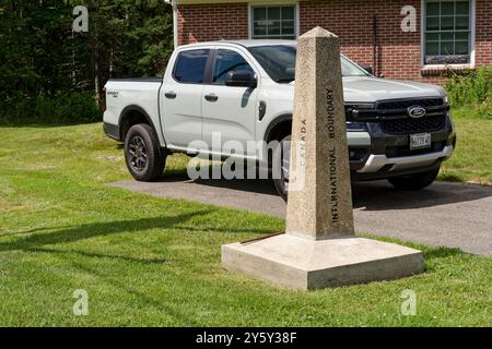 Saint-Augustin-de-Woburn, Quebec, Kanada - 19. Juli 2024: Ein Stein markiert die Grenze zwischen Kanada und den USA, dahinter ein amerikanischer LKW und ein Zollamt. Stockfoto