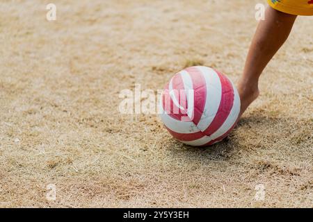 Eine Nahaufnahme eines Kindes, das einen rot-weiß gestreiften Ball auf einem trockenen, grasbewachsenen Feld tritt. Die sonnige Outdoor-Szene vermittelt Verspieltheit und Freizeit. Stockfoto