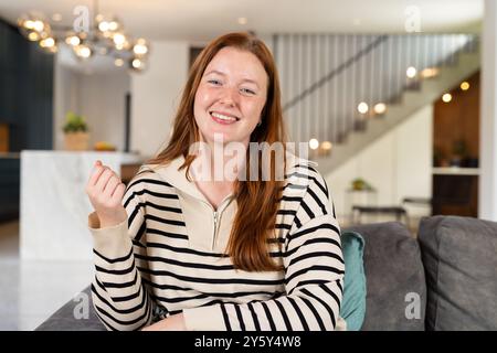 Lächelnde Frau in gestreiftem Pullover, die auf der Couch im modernen Wohnzimmer sitzt, auf Videoanruf Stockfoto