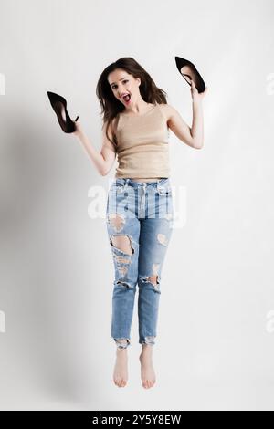 Lebendiges Bild einer verspielten jungen Frau, die fröhlich springt und ihre High Heels hält. Sie trägt zerrissene Jeans und ein glitzerndes Oberteil, das eine sorglose, energisch zeigt Stockfoto