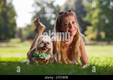 Eine lächelnde Frau entspannt sich im Gras eines sonnendurchfluteten Parks neben ihrem süßen Shih Tzu-Hund in einem bunten Outfit. Stockfoto