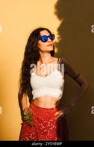 Modische junge Frau mit lockigem Haar in Sonnenbrille, die sich an einem hellen Sommertag selbstbewusst vor einer leuchtend gelben Wand posiert. Stockfoto