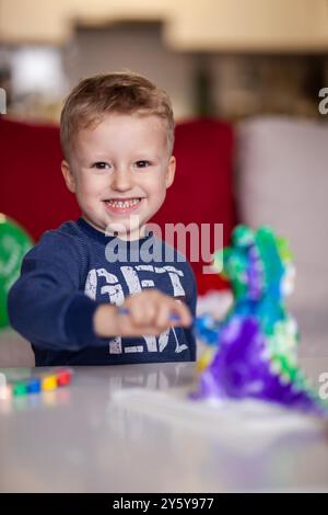 Ein fröhliches kleines Kind, das breit lächelt, während es mit lebhaften Dinosaurierfiguren auf einem Tisch in gemütlicher Atmosphäre spielt. Stockfoto