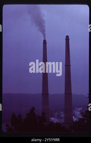 Zwei hoch aufragende Schornsteine erheben sich in den Abendhimmel und emittieren Rauchwolken aus einem Industriewerk im Mittleren Westen, wahrscheinlich in Toledo, Ohio, um die 1970er Jahre Das Bild verdeutlicht die Abhängigkeit der Region von der Schwerindustrie in diesem Zeitraum, was sowohl zum Wirtschaftswachstum als auch zu ökologischen Herausforderungen beiträgt. Stockfoto