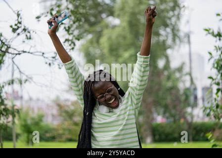 Die afroamerikanische Frau hebt fröhlich ihre Arme, während sie ihr Smartphone hält, und drückt ihre Begeisterung im grünen Park aus. Stockfoto