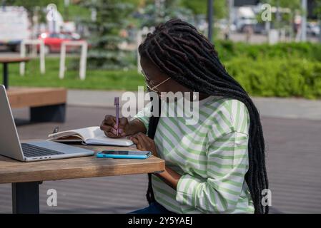 Afroamerikanerin sitzt draußen an einem Tisch, schreibt in einem Notizbuch mit einem Laptop neben ihr und genießt eine produktive Lerneinheit. Stockfoto