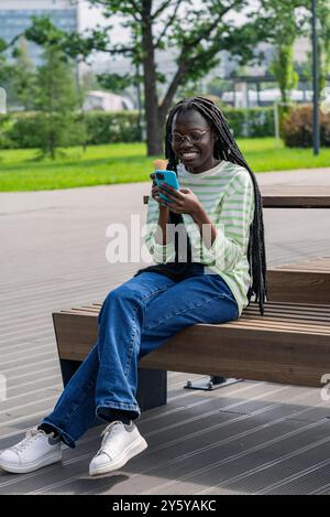 Die junge afroamerikanische Frau sitzt auf einer Bank, lächelt, während sie Eis isst und ihr Smartphone benutzt, um einen sonnigen Tag im Freien zu genießen. Stockfoto