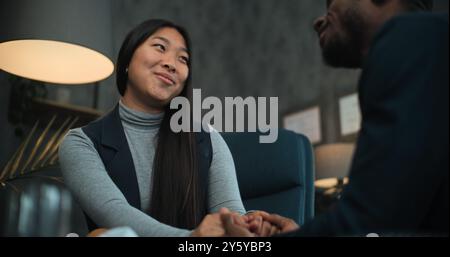 Asiatische Frau lächelt, spricht mit ihrem Freund oder Ehemann während der Beratung mit dem Therapeuten. Glückliches multiethnisches Paar sitzt auf Stühlen und hält sich an den Händen, findet Kompromisse nach Streit über die Paartherapie. Stockfoto