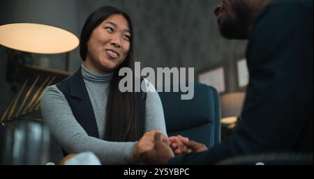 Asiatische Frau lächelt, spricht mit ihrem Freund oder Ehemann während der Beratung mit dem Therapeuten. Glückliches multiethnisches Paar sitzt auf Stühlen und hält sich an den Händen, findet Kompromisse nach Streit über die Paartherapie. Stockfoto