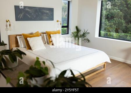 Modernes Schlafzimmer mit weißer Bettwäsche, gelben Kissen, großen Fenstern mit Blick auf das Grün, Kopierraum Stockfoto