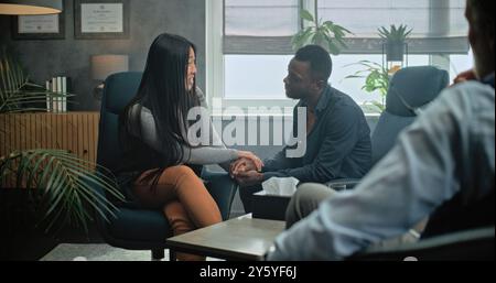 Glückliches multiethnisches Paar sitzt Hand in Hand auf Stühlen, lacht, sieht sich an, findet Kompromisse nach Kampf gegen Paartherapie. Junge Familie auf erfolgreicher Beratung mit Psychologin. Stockfoto