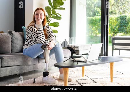 Sitzend auf der Couch, lächelnde Frau mit einer Beinprothese, mit Laptop und Mikrofon Stockfoto