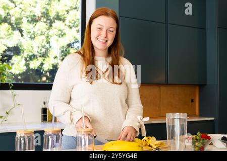 Lächelnde Frau, die Obst-Smoothie in moderner Küche mit frischen Zutaten zubereitet Stockfoto