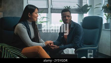 Glückliches multiethnisches Ehepaar in Beratungssitzung mit Therapeutin. Afroamerikanischer Mann und asiatische Frau sitzen auf Stühlen und halten Hände, nachdem sie sich über die Paartherapie gestritten haben. Psychologe spricht mit der jungen Familie. Stockfoto
