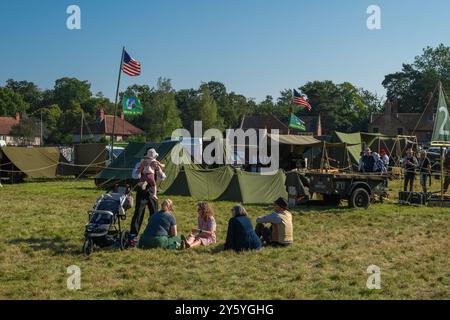 Holt, Norfolk, Vereinigtes Königreich - 21. SEPTEMBER 2024: Familie in Vintage-Kleidung mit Baby sitzt auf dem Rasen vor Militärzelten und amerikanischer Flagge Stockfoto