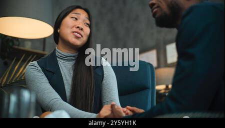 Asiatische Frau lächelt, spricht mit ihrem Freund oder Ehemann während der Beratung mit dem Therapeuten. Glückliches multiethnisches Paar sitzt auf Stühlen und hält sich an den Händen, findet Kompromisse nach Streit über die Paartherapie. Stockfoto