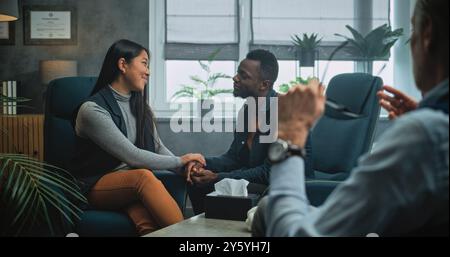 Glückliches multiethnisches Paar sitzt Hand in Hand auf Stühlen, lacht, sieht sich an, findet Kompromisse nach Kampf gegen Paartherapie. Junge Familie auf erfolgreicher Beratung mit Psychologin. Stockfoto