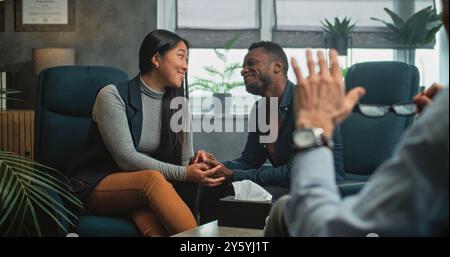 Glückliches multiethnisches Paar sitzt Hand in Hand auf Stühlen, lacht, sieht sich an, findet Kompromisse nach Kampf gegen Paartherapie. Junge Familie auf erfolgreicher Beratung mit Psychologin. Stockfoto