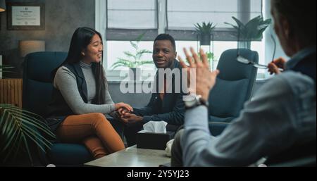 Glückliches multiethnisches Paar sitzt Hand in Hand auf Stühlen, lacht, sieht sich an, findet Kompromisse nach Kampf gegen Paartherapie. Junge Familie auf erfolgreicher Beratung mit Psychologin. Stockfoto