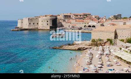 Besucher genießen den weichen Sand und das pulsierende blaue Wasser des Strandes von Dubrovnik, während historische Gebäude majestätisch im Hintergrund stehen und Kroatiens reiches kulturelles Erbe unter der Sonne zeigen. Stockfoto