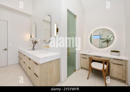Modernes Bad mit hellen Holzschränken, großen Spiegeln und eleganter Einrichtung mit einem Kosmetikbereich mit rundem beleuchtetem Spiegel. Stockfoto