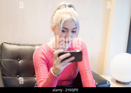 Eine junge Frau in einem rosafarbenen Pullover konzentriert sich intensiv auf ihr Smartphone und zeigt einen konzentrierten Ausdruck in einer gemütlichen Innenumgebung mit sanfter Beleuchtung Stockfoto
